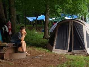 A male Camper sitting alone in the front of his camp tent watching other people play.